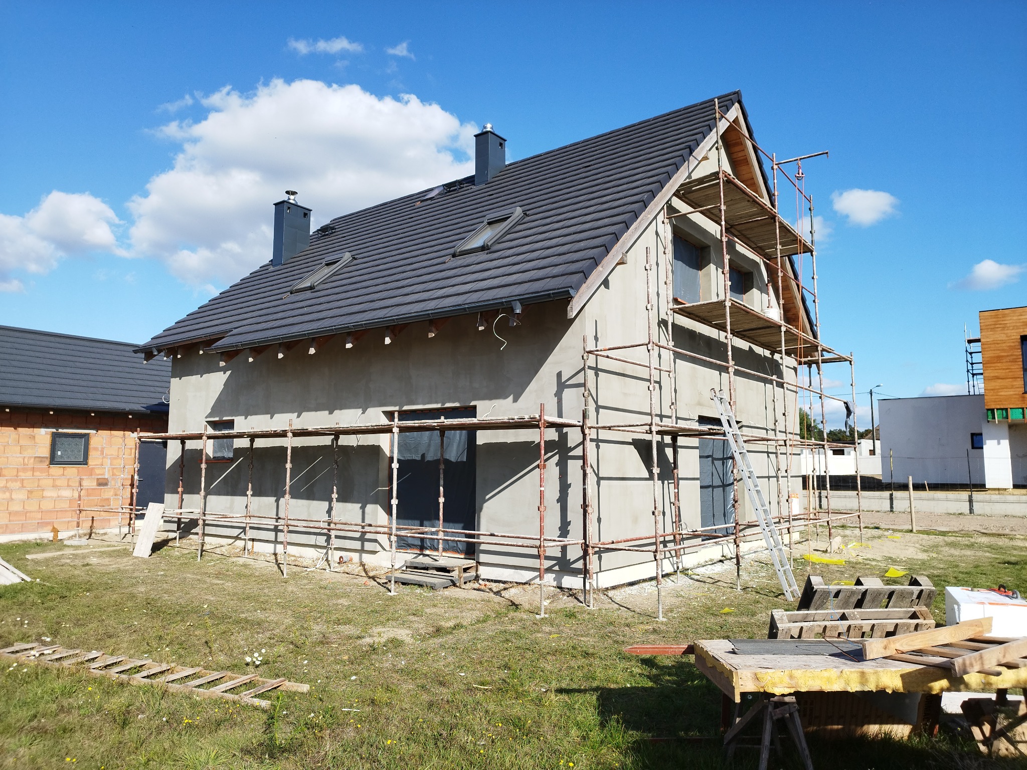 Dom jednorodzinny w trakcie ocieplania, widoczne rusztowania, nieotynkowana elewacja w kolorze szarym, dach pokryty ciemną dachówką, błękitne niebo z chmurami w tle.