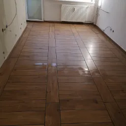 Ułożona podłoga z brązowych płytek imitujących drewno, z szerokimi pasami płytek wzdłuż ścian i w poprzek, widok na balkon i grzejnik, instalacje elektryczne wystające ze ścian.