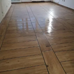 Ułożona podłoga z płytek imitujących drewno w jasnym pomieszczeniu, widoczne fugi i odbicie światła na powierzchni płytek, grzejnik na ścianie w tle.