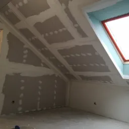 Poddasze w trakcie prac wykończeniowych, widoczne płyty kartonowo-gipsowe na skosach dachu, szpachlowane łączenia, okno dachowe z czerwonym obramowaniem, wiadro z narzędziami na podłodze.