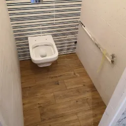 Remontowana łazienka: podwieszana toaleta z chromowanym przyciskiem spłukiwania na ścianie w biało-granatowe pasy, na podłodze panele imitujące drewno, widoczne niedokończone prace glazurnicze...