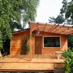 Drewniany domek z tarasem i pergolą, oświetlony wiszącą lampą, otoczony zielenią drzew i krzewów.