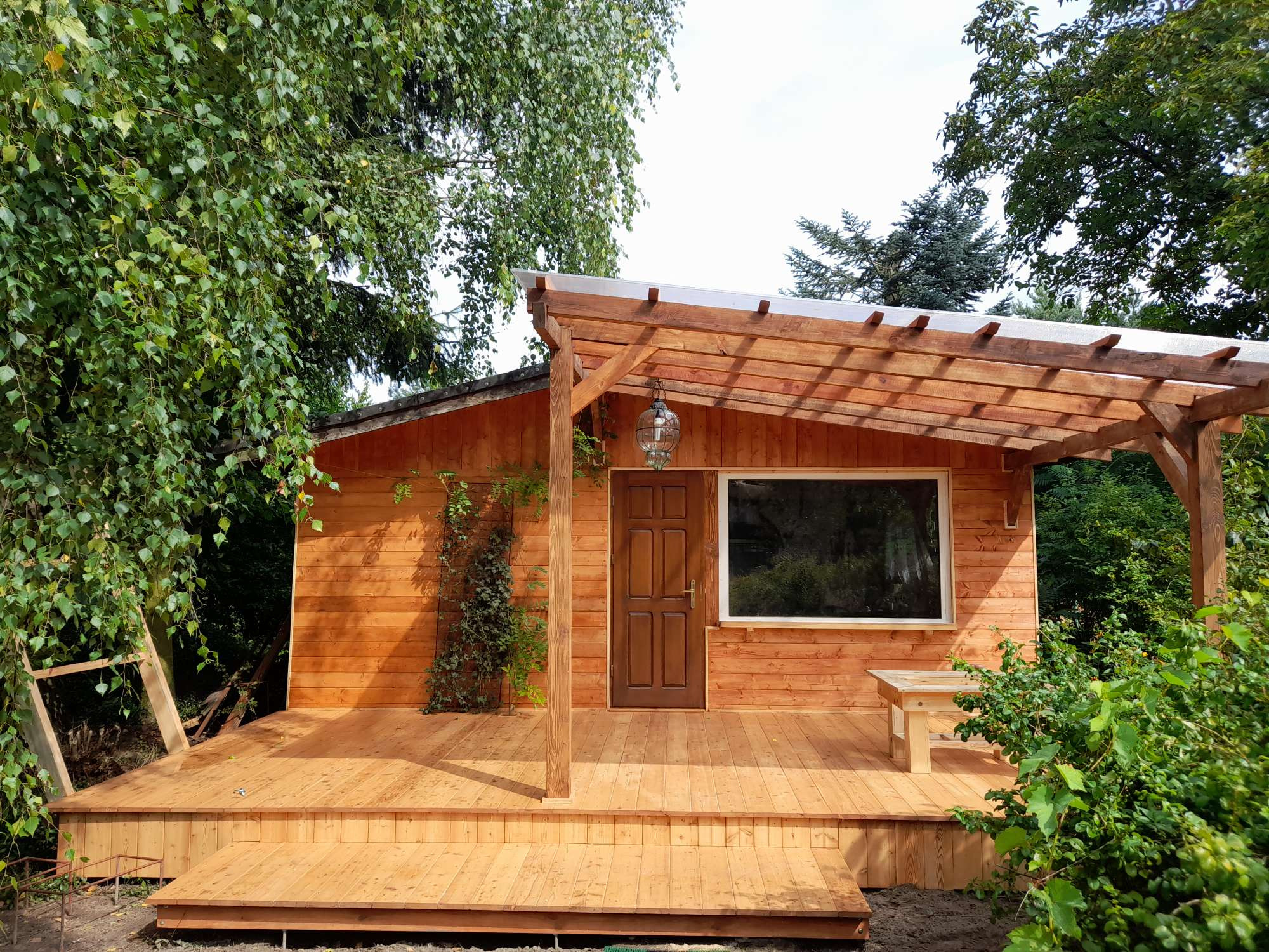 Drewniany domek z tarasem i pergolą, oświetlony wiszącą lampą, otoczony zielenią drzew i krzewów.