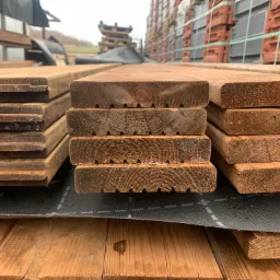 Stos desek tarasowych z frezowanymi rowkami na spodniej stronie, ułożonych na czarnej macie, widoczne cegły w tle.