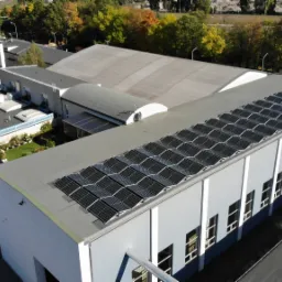 Widok z góry na budynek przemysłowy z zainstalowanymi panelami słonecznymi na dachu, otoczony zielenią i parkingiem z samochodami.