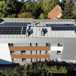 Soltech Group Sp. z o.o. - Z lotu ptaka, nowoczesny budynek z szaro-brązową elewacją i napisem 'ENERGOTHERM', na którego płaskim dachu zamontowano trzy rzędy czarnych modułów fotowoltaicznych. W tle droga i zieleń.