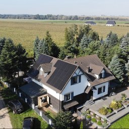 Soltech Group Sp. z o.o. - Z lotu ptaka: dom jednorodzinny z białymi i ciemnymi elewacjami, z licznymi ciemnymi panelami słonecznymi na wielospadowym dachu. Wokół zielone drzewa i rozległe pola uprawne.
