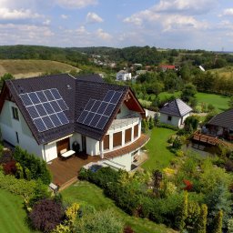 Soltech Group Sp. z o.o. - Nowoczesny dom z dwoma zestawami paneli słonecznych na ciemnym dachu, z okrągłym balkonem i drewnianym tarasem. Rozległy, zielony ogród z oczkiem wodnym i altaną, ujęcie z lotu ptaka.