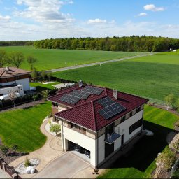 Soltech Group Sp. z o.o. - Z lotu ptaka: nowoczesny dom z bordowym dachem pokrytym panelami fotowoltaicznymi, otoczony zielonym trawnikiem i polami uprawnymi, w tle las.