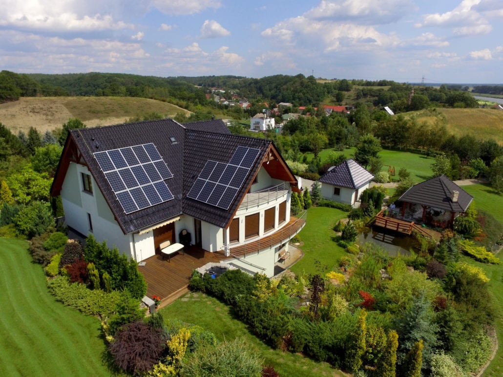 Dom jednorodzinny z panelami słonecznymi na dachu, widok z lotu ptaka na zadbany ogród z oczkiem wodnym i dwoma budynkami gospodarczymi.