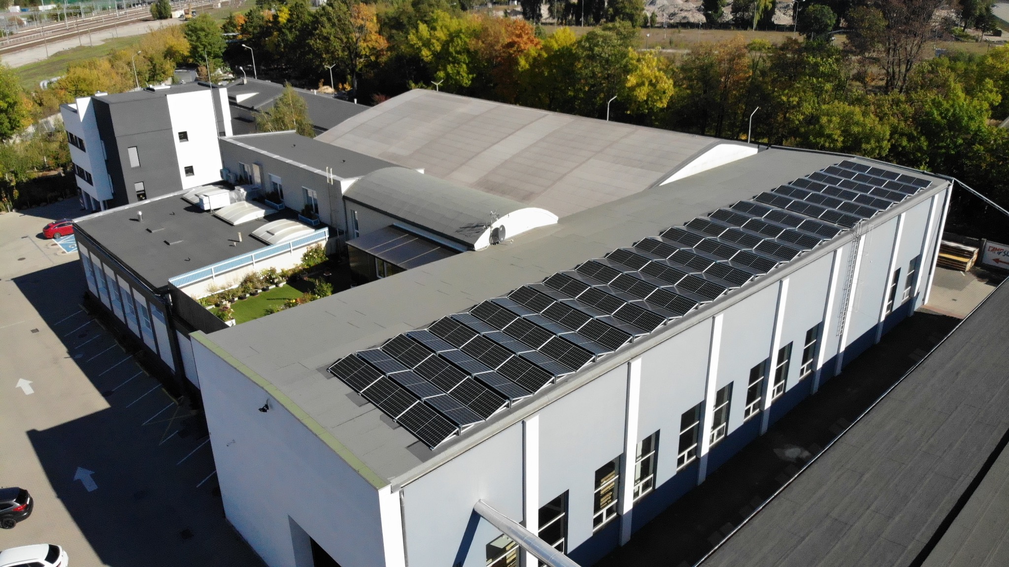 Widok z góry na budynek przemysłowy z zainstalowanymi panelami słonecznymi na dachu, otoczony zielenią i parkingiem z samochodami.