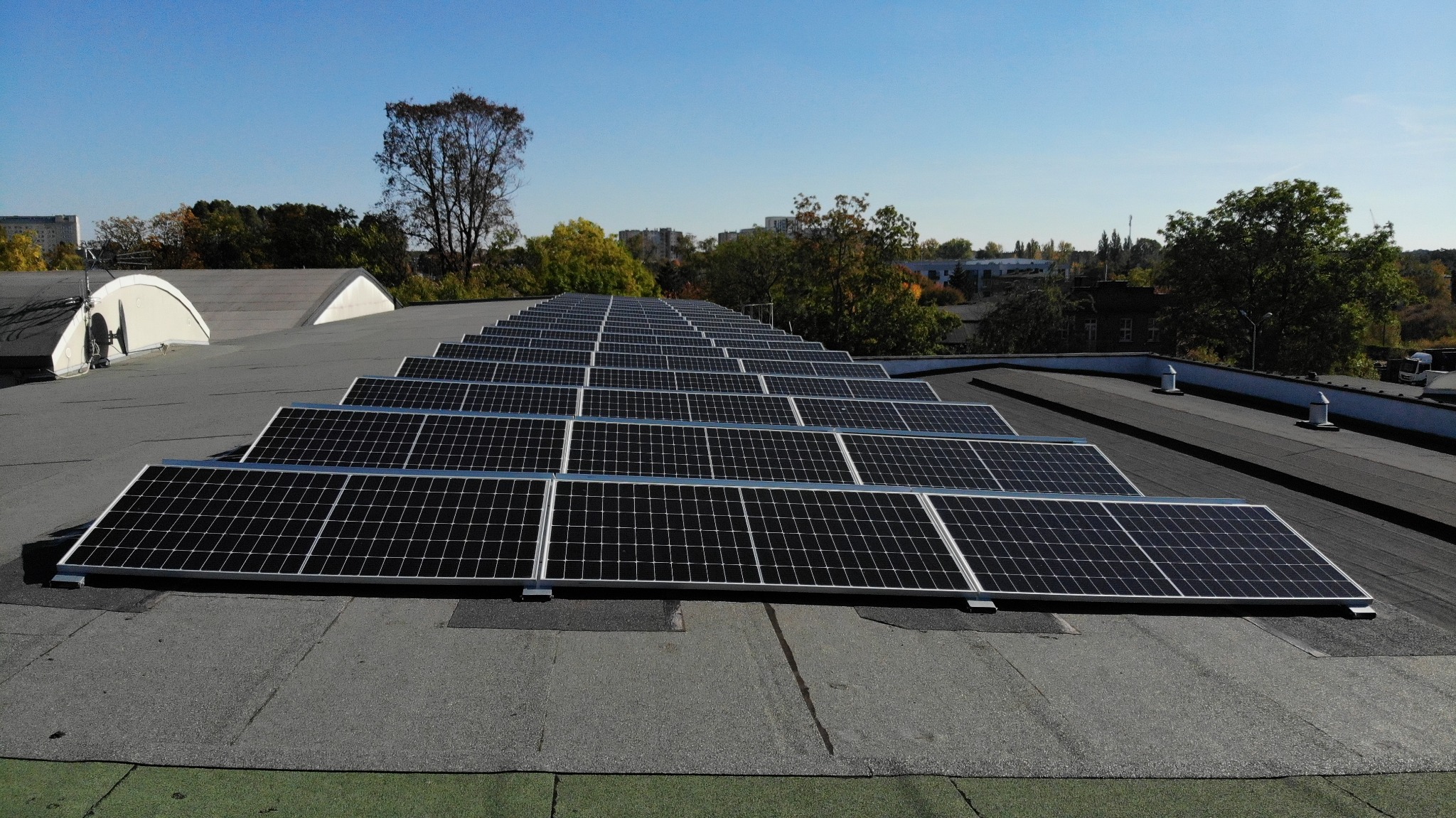Rząd paneli fotowoltaicznych na płaskim dachu budynku w słoneczny dzień. Widoczne drzewa i zabudowa miejska w tle. Instalacja wykonana z dbałością o detale.