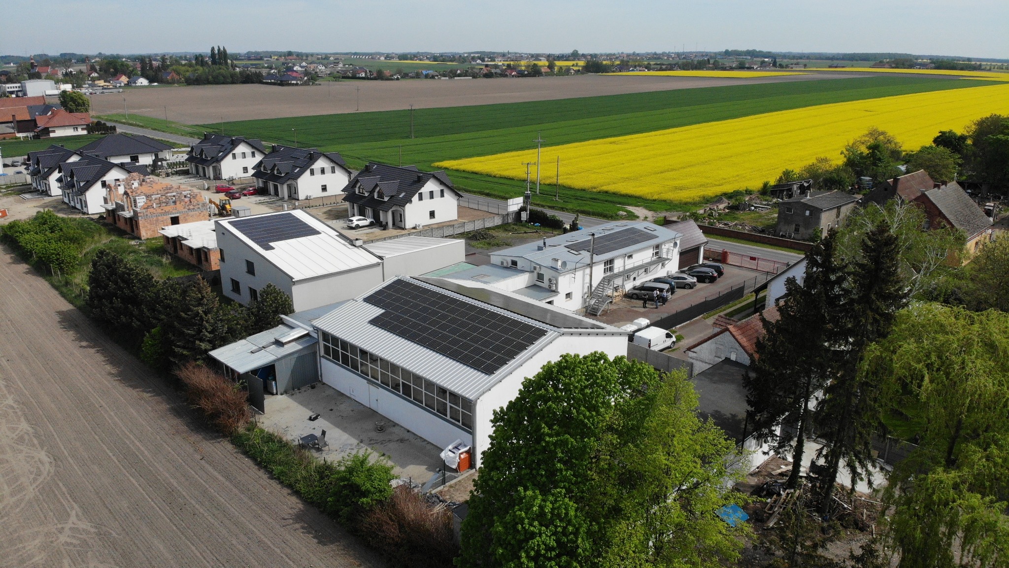 Widok z drona na budynki z panelami słonecznymi na dachach, w tle pola uprawne i zabudowa wiejska. Panele zamontowane na dachu budynku z szarym dachem.