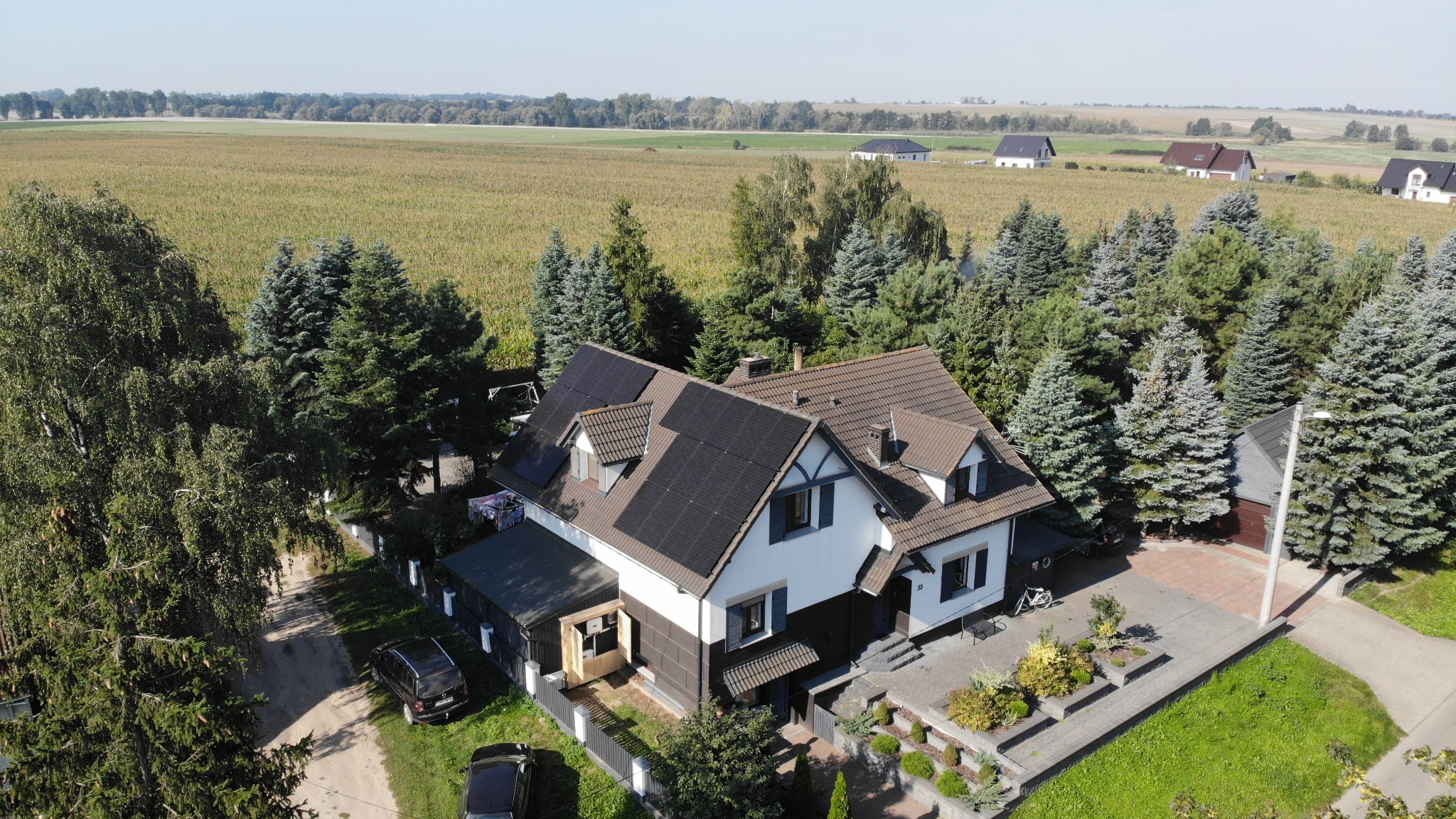 Dom jednorodzinny z panelami fotowoltaicznymi na dachu, otoczony zielenią i polami, widziany z lotu ptaka w słoneczny dzień. Widoczne detale architektoniczne i starannie zagospodarowany ogród.