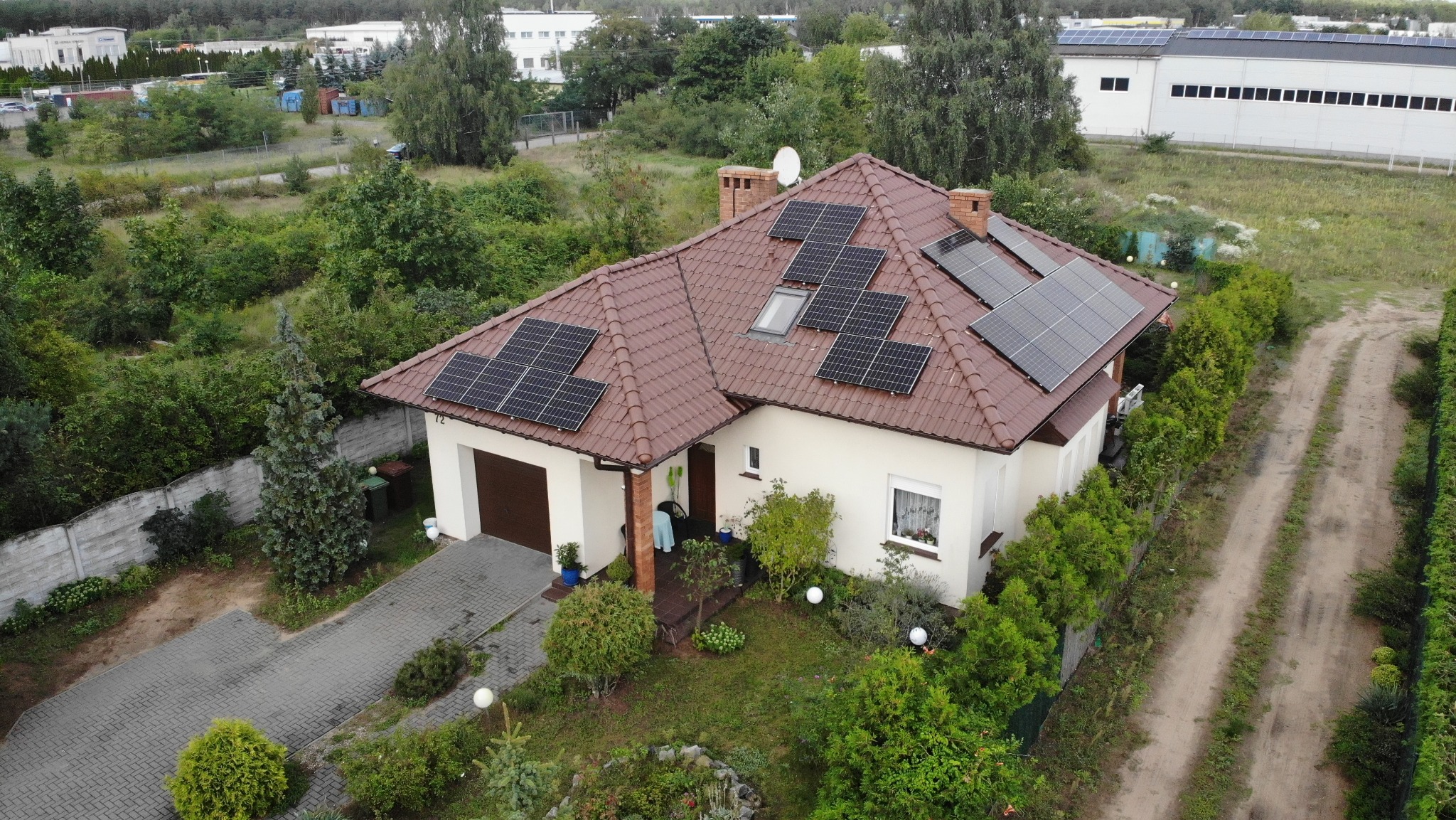 Dom jednorodzinny z brązową dachówką i panelami słonecznymi na dachu, widok z góry, otoczony zielenią. Na dachu widoczne okno dachowe.