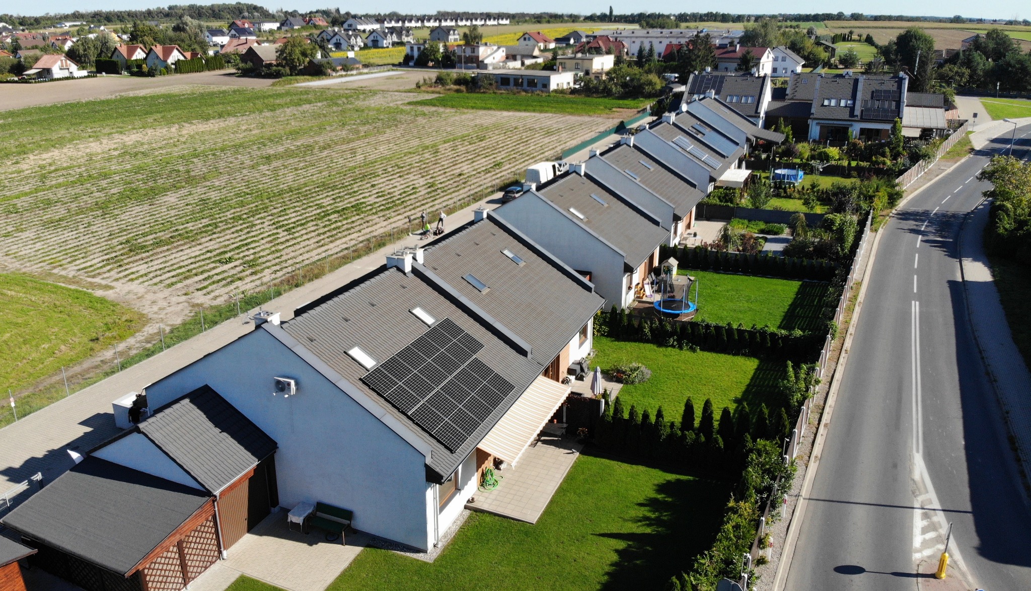 Widok z góry na szereg domów z panelami słonecznymi na dachach, otoczonych zielonymi ogrodami i polami uprawnymi w tle. Słoneczny dzień.