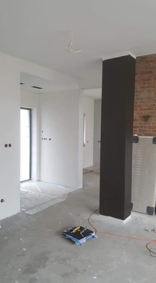Wnętrze w trakcie prac wykończeniowych: świeżo pomalowane na biało ściany, czarny filar, zabezpieczona folią podłoga, widoczne narzędzia malarskie, fragment odsłoniętej ceglanej ściany.