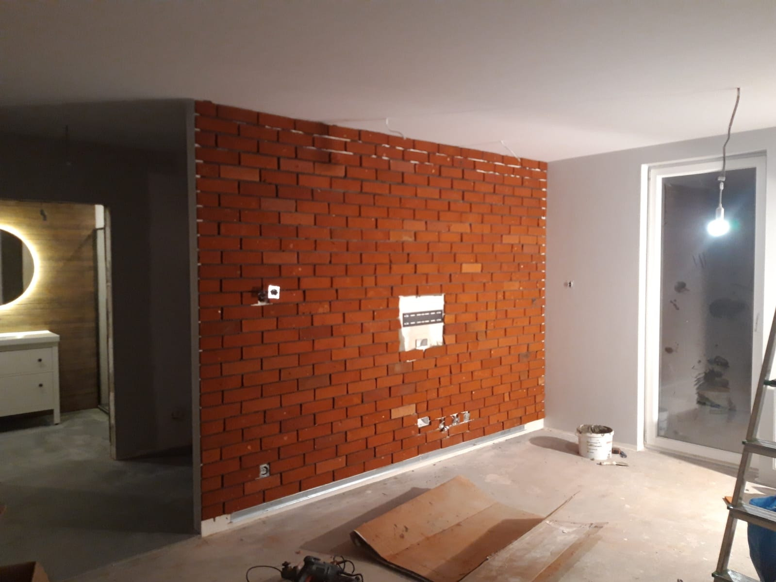 Montaż dekoracyjnej ściany z cegły w trakcie remontu, widoczne puszki elektryczne i częściowo wykończona instalacja, narzędzia na podłodze, wnętrze łazienki z okrągłym lustrem w tle.