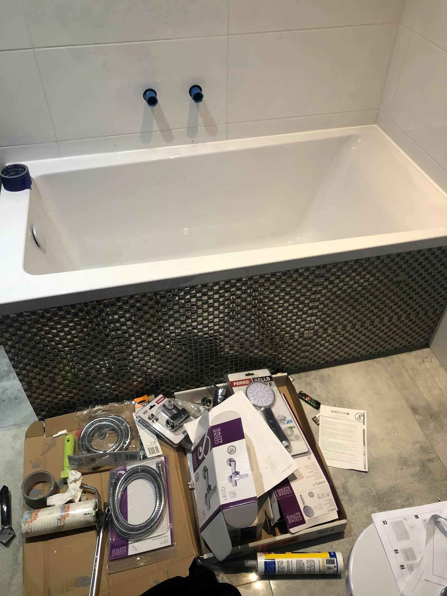 Biała wanna z mozaikową obudową w trakcie montażu, widoczne niepodłączone rury, kartony z armaturą i narzędzia na podłodze.