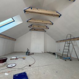 Solid Bud - Poddasze w trakcie malowania na biało, zabezpieczone drewniane elementy, drabina, narzędzia malarskie, okno dachowe. Widok na surowe wnętrze przed wykończeniem.