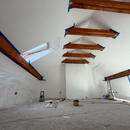 Solid Bud - Białe poddasze ze skośnym sufitem w trakcie malowania. Widoczne drewniane belki zabezpieczone taśmą, okno dachowe i narzędzia malarskie.