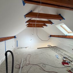 Solid Bud - Poddasze w trakcie malowania: drewniane belki zabezpieczone taśmą, ściany pokryte świeżą, białą farbą. Widoczne narzędzia malarskie i zabezpieczenia podłogi folią.