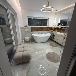 Solid Bud - Elegancka łazienka z wolnostojącą wanną, beżowymi płytkami imitującymi marmur i okrągłym, złotym oświetleniem. Widoczny puf i toaleta. Całość w jasnych, ciepłych barwach.