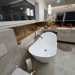 Solid Bud - Nowoczesna łazienka z wolnostojącą wanną, marmurowymi płytkami i złotymi akcentami. Widoczne okno, toaleta i umywalka z szafkami. Oświetlenie punktowe i dekoracje.