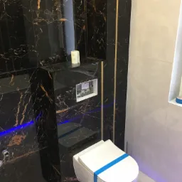 Elegancka toaleta podwieszana z czarną marmurową obudową, wykończona złotymi akcentami. Na blacie rolka papieru toaletowego. Biała podłoga z marmurowym wzorem.