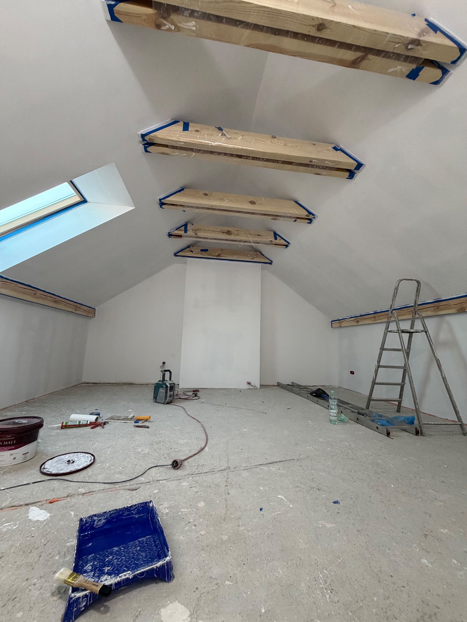 Poddasze w trakcie malowania na biało, zabezpieczone drewniane elementy, drabina, narzędzia malarskie, okno dachowe. Widok na surowe wnętrze przed wykończeniem.