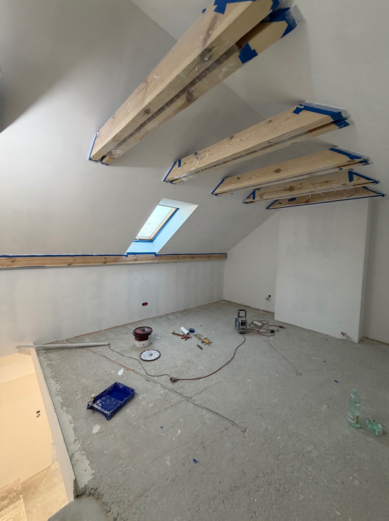 Poddasze w trakcie prac wykończeniowych: zabezpieczone drewniane belki stropowe, okno dachowe, świeżo pomalowane ściany, narzędzia i akcesoria malarskie na podłodze.