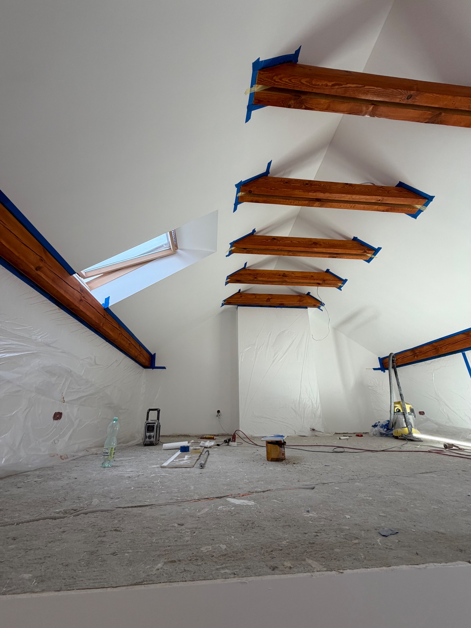 Białe poddasze ze skośnym sufitem w trakcie malowania. Widoczne drewniane belki zabezpieczone taśmą, okno dachowe i narzędzia malarskie.