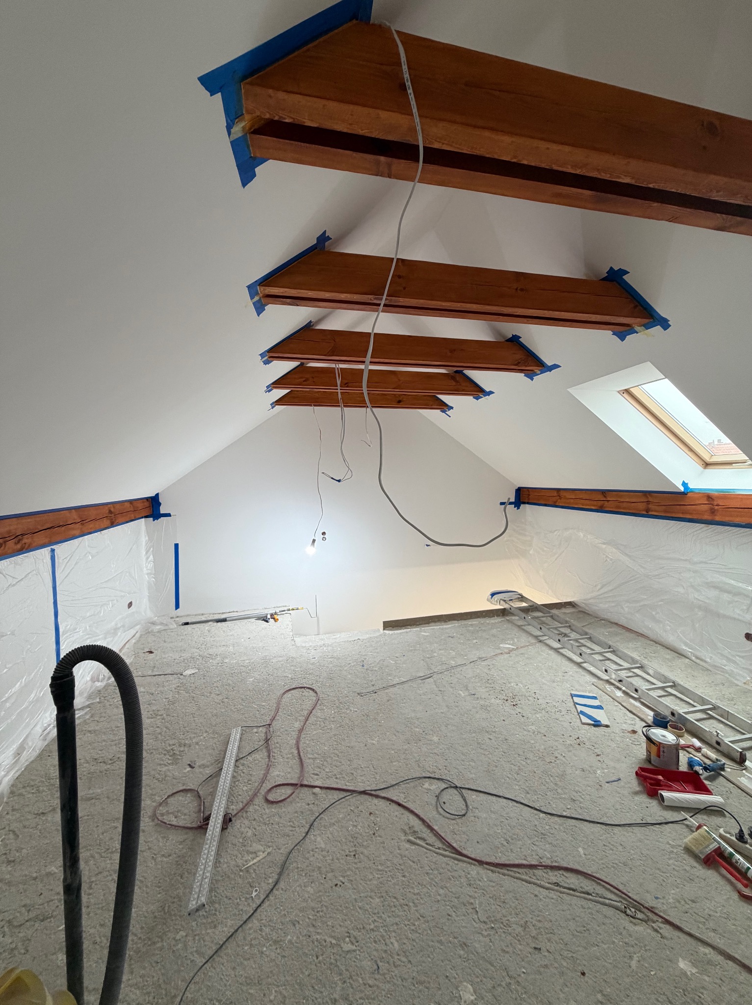 Poddasze w trakcie malowania: drewniane belki zabezpieczone taśmą, ściany pokryte świeżą, białą farbą. Widoczne narzędzia malarskie i zabezpieczenia podłogi folią.