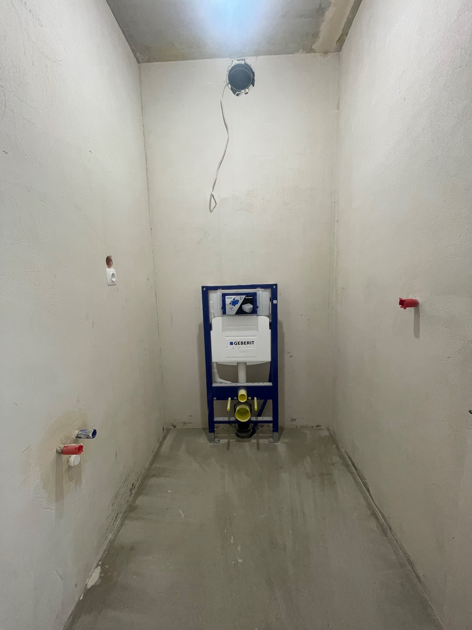 Stelaż podtynkowy Geberit w surowej łazience przed wykończeniem. Widoczne wystające rury i instalacja elektryczna. Ściany w stanie deweloperskim, surowy beton na podłodze.