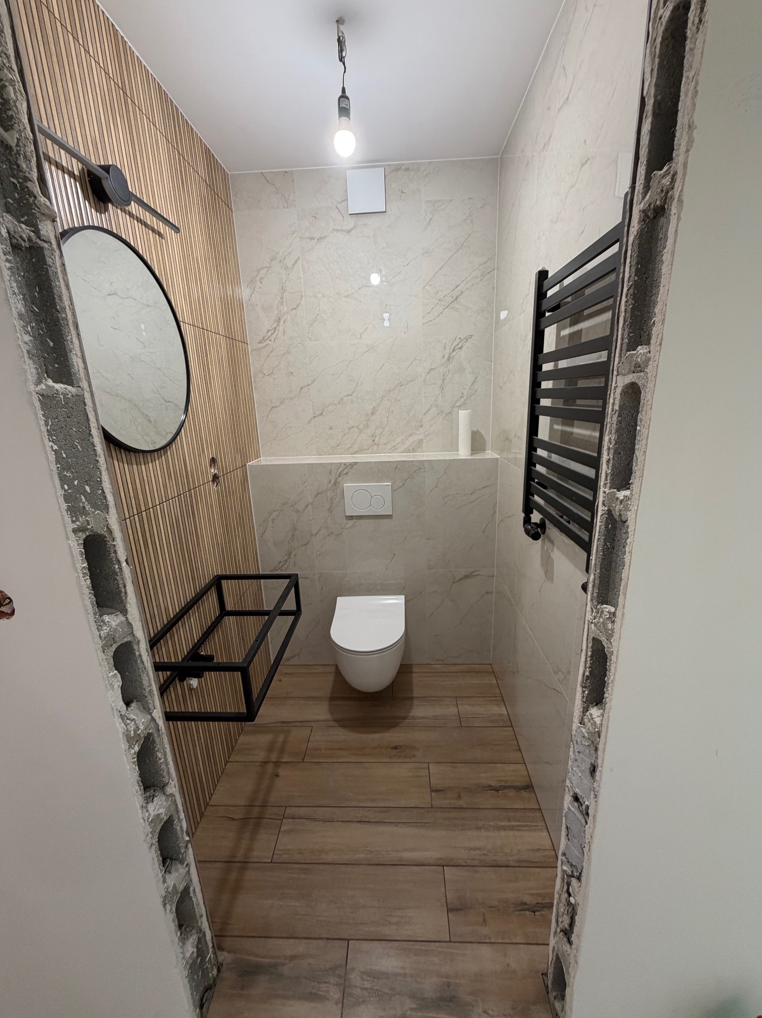Nowoczesna łazienka z drewnopodobną podłogą i ścianą z lamelami, widok przez niedokończone ościeżnice. Grzejnik drabinkowy i okrągłe lustro.