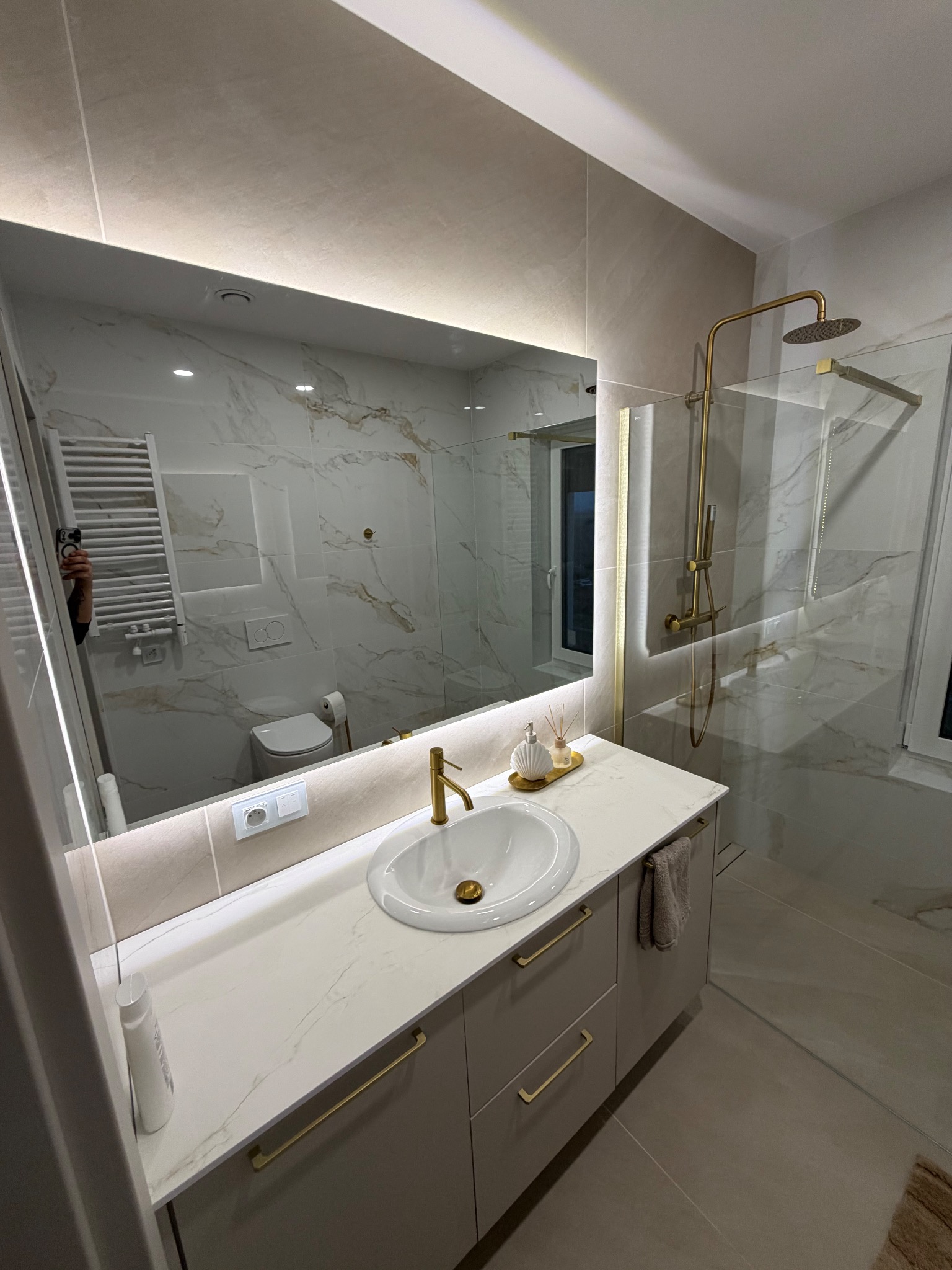 Elegancka łazienka z marmurowymi płytkami, złotą armaturą i podświetlanym lustrem. Umywalka nablatowa, kabina prysznicowa typu walk-in. Nowoczesny design.