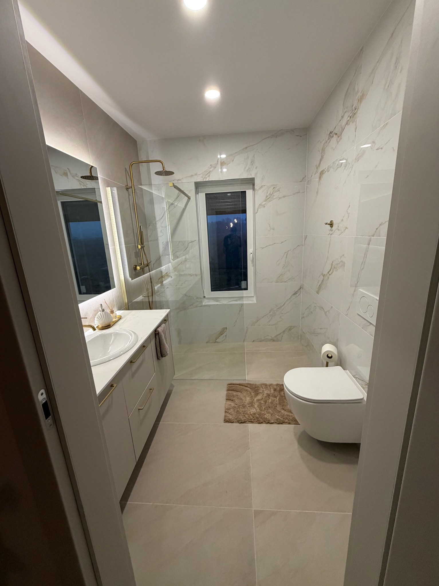 Elegancka łazienka z kabiną prysznicową typu walk-in, umywalką z szafką w odcieniach szarości i toaletą podwieszaną. Płytki imitujące marmur na ścianach i podłodze.