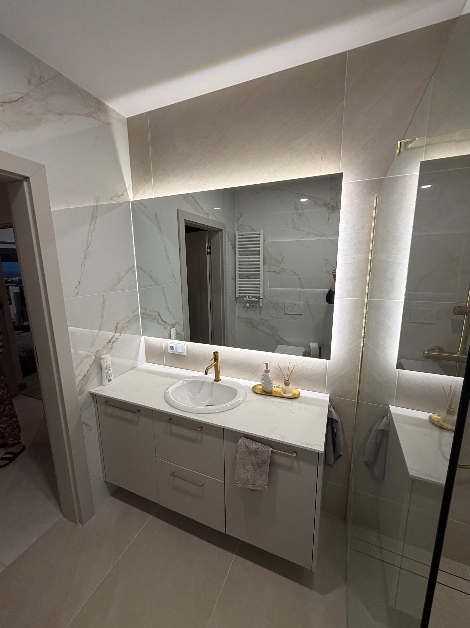 Elegancka łazienka z podświetlanym lustrem, umywalką nablatową i szafką w odcieniach beżu. Marmurowe płytki na ścianach i podłodze. Widoczne akcesoria łazienkowe.