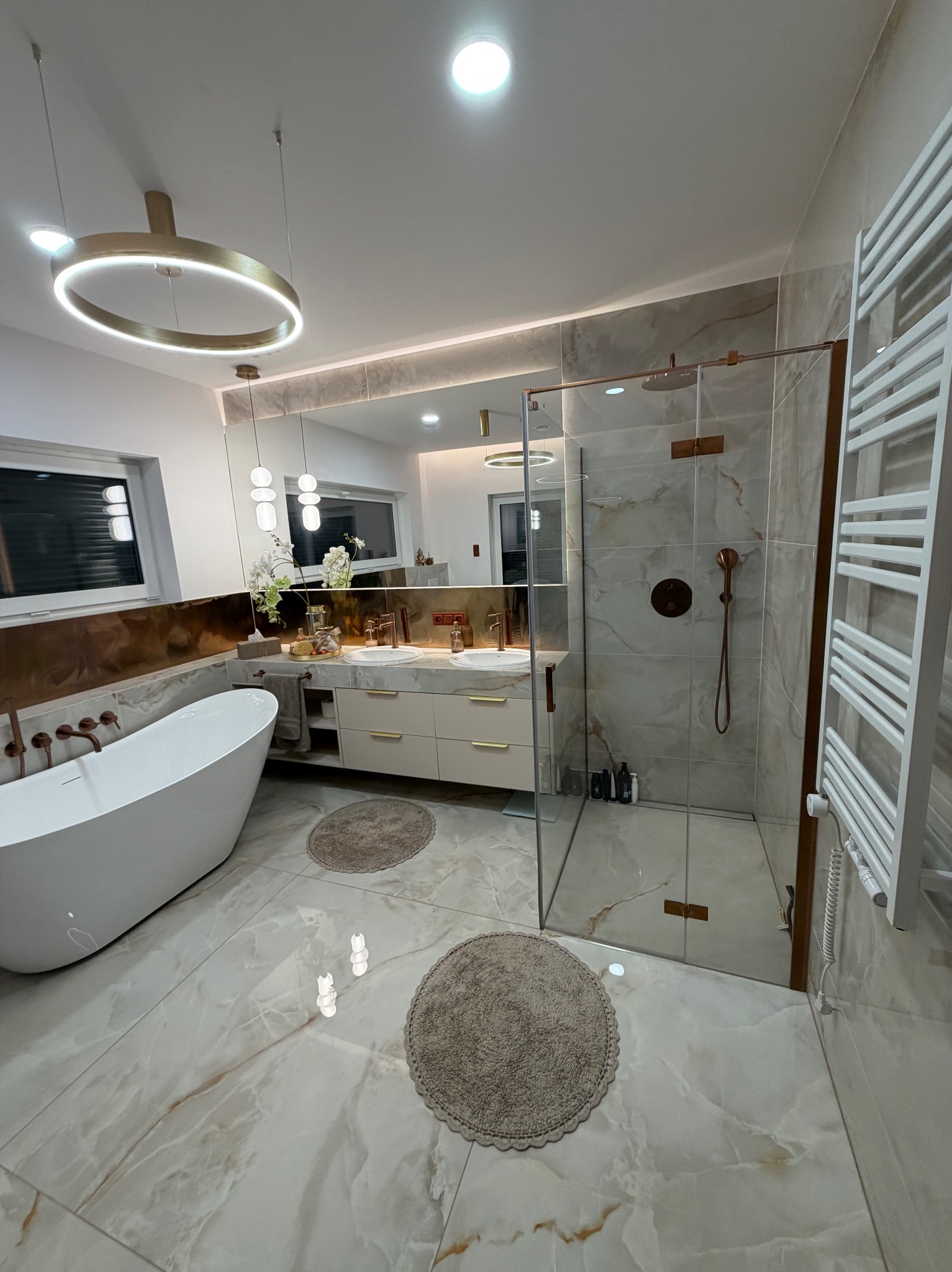 Elegancka łazienka z wanną wolnostojącą, kabiną prysznicową ze szklanymi drzwiami i marmurowymi płytkami. Złote akcenty armatury i oświetlenia dodają luksusowego charakteru.