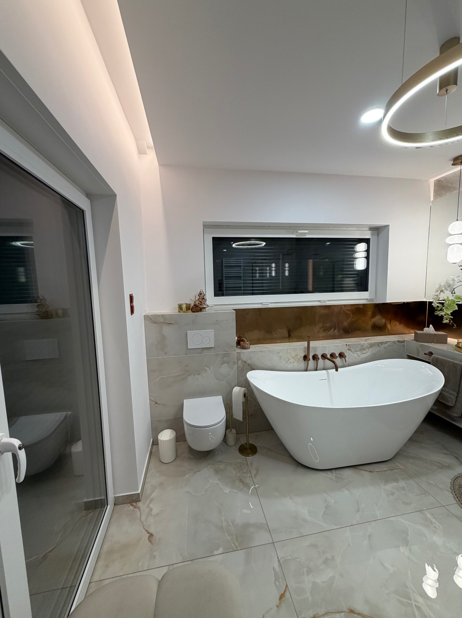 Nowoczesna łazienka z wolnostojącą wanną, marmurową podłogą i złotymi akcentami. Okno z żaluzjami, designerska lampa sufitowa. Widok z perspektywy drzwi kabiny prysznicowej.