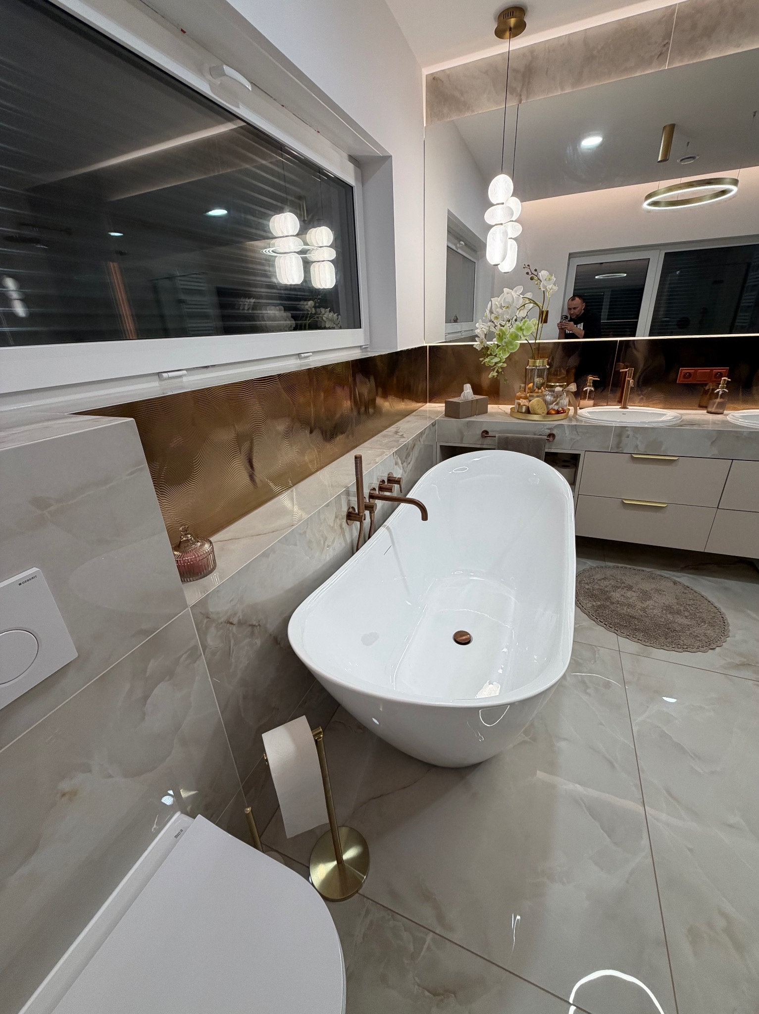 Nowoczesna łazienka z wolnostojącą wanną, marmurowymi płytkami i złotymi akcentami. Widoczne okno, toaleta i umywalka z szafkami. Oświetlenie punktowe i dekoracje.