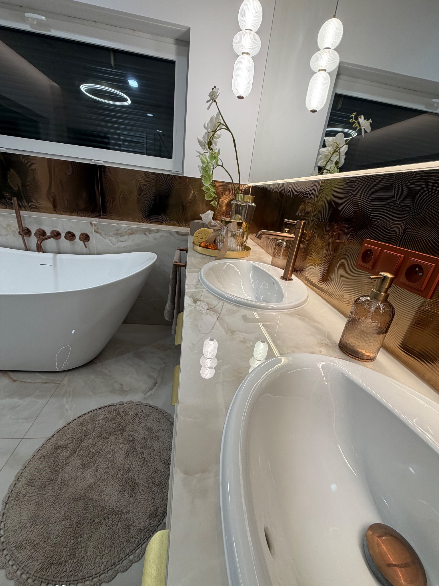 Nowoczesna łazienka z wolnostojącą wanną, dwiema umywalkami nablatowymi i złotymi akcentami. Widoczne płytki imitujące marmur i dekoracyjne oświetlenie.