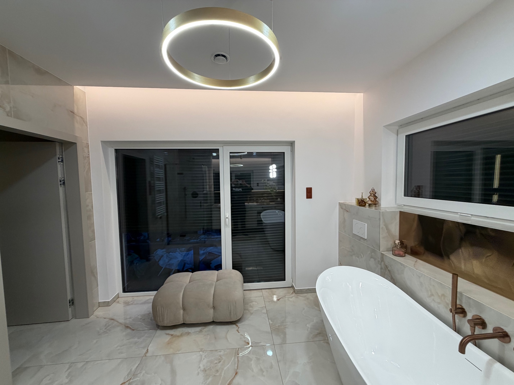 Nowoczesna łazienka z marmurową podłogą, wolnostojącą wanną i oknem balkonowym. Okrągła lampa sufitowa dodaje elegancji. Widoczny puf i złote akcenty.