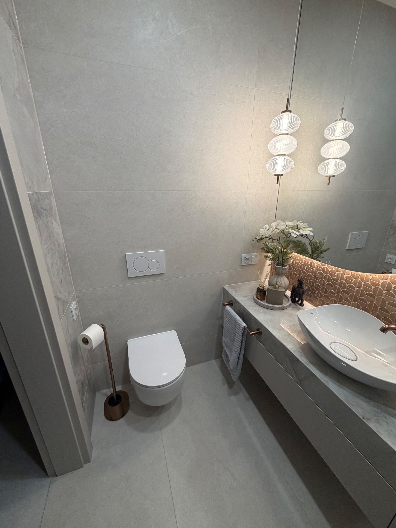 Nowoczesna łazienka w szarościach z wiszącą toaletą, umywalką nablatową i dekoracyjnym oświetleniem. Uwagę zwraca lustro z podświetleniem i nietypowy kształt lamp.