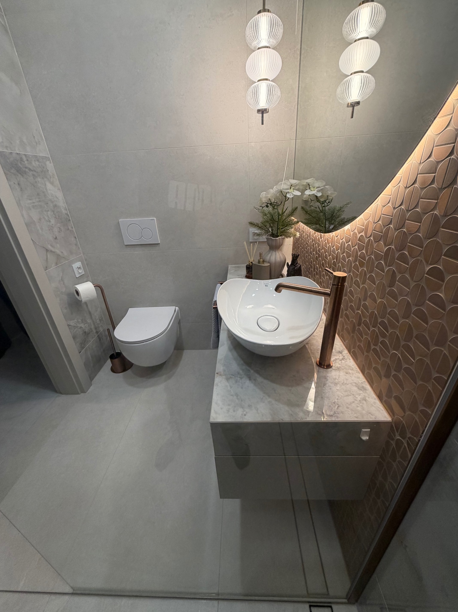 Nowoczesna łazienka z umywalką nablatową, mozaiką w odcieniach brązu i szarości oraz wiszącą toaletą. Całość dopełnia designerskie oświetlenie.