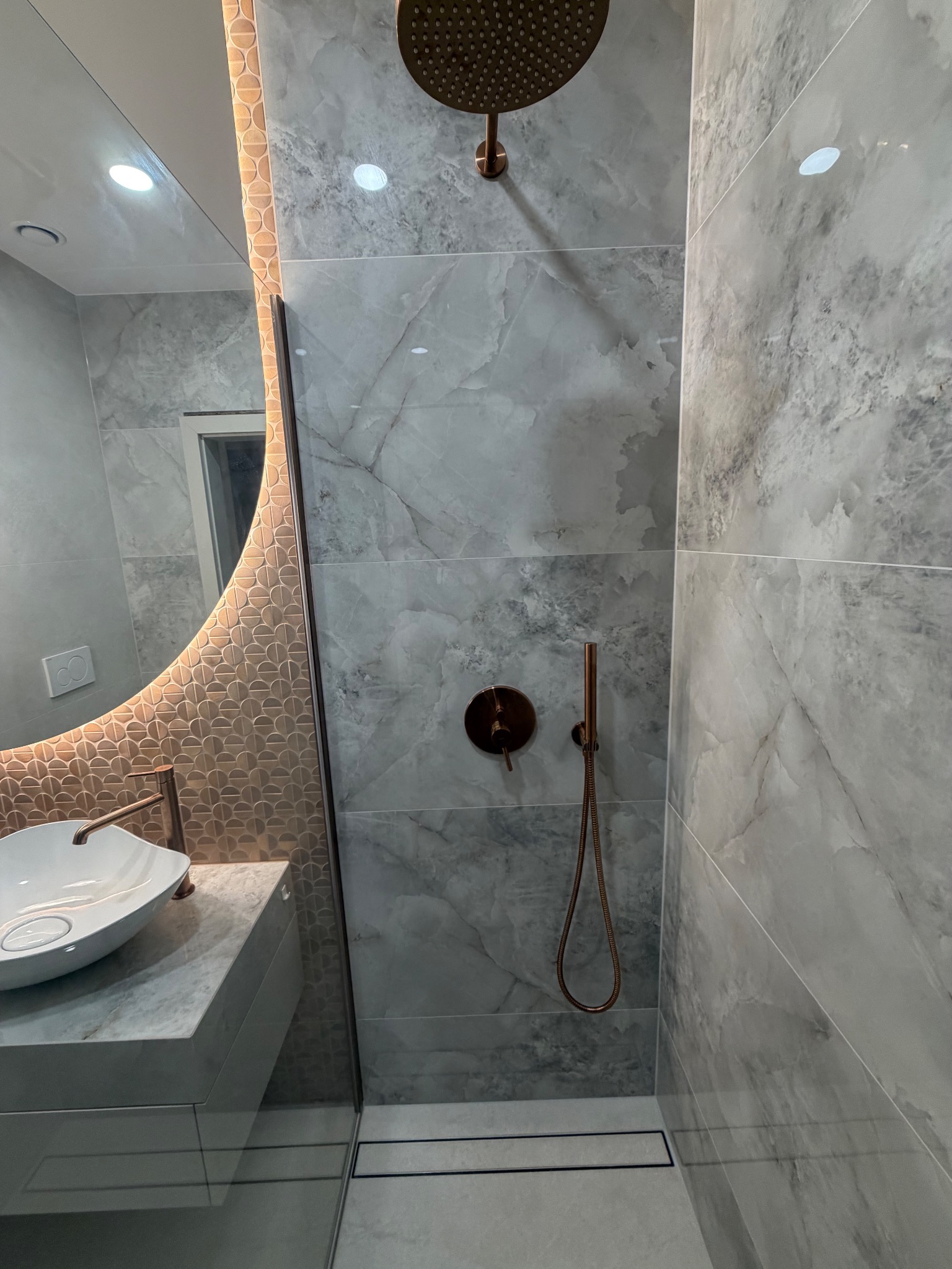 Nowoczesna łazienka z szarymi płytkami imitującymi marmur, deszczownicą w kolorze miedzi i okrągłym lustrem z podświetleniem. Minimalistyczny design, eleganckie wykończenie.