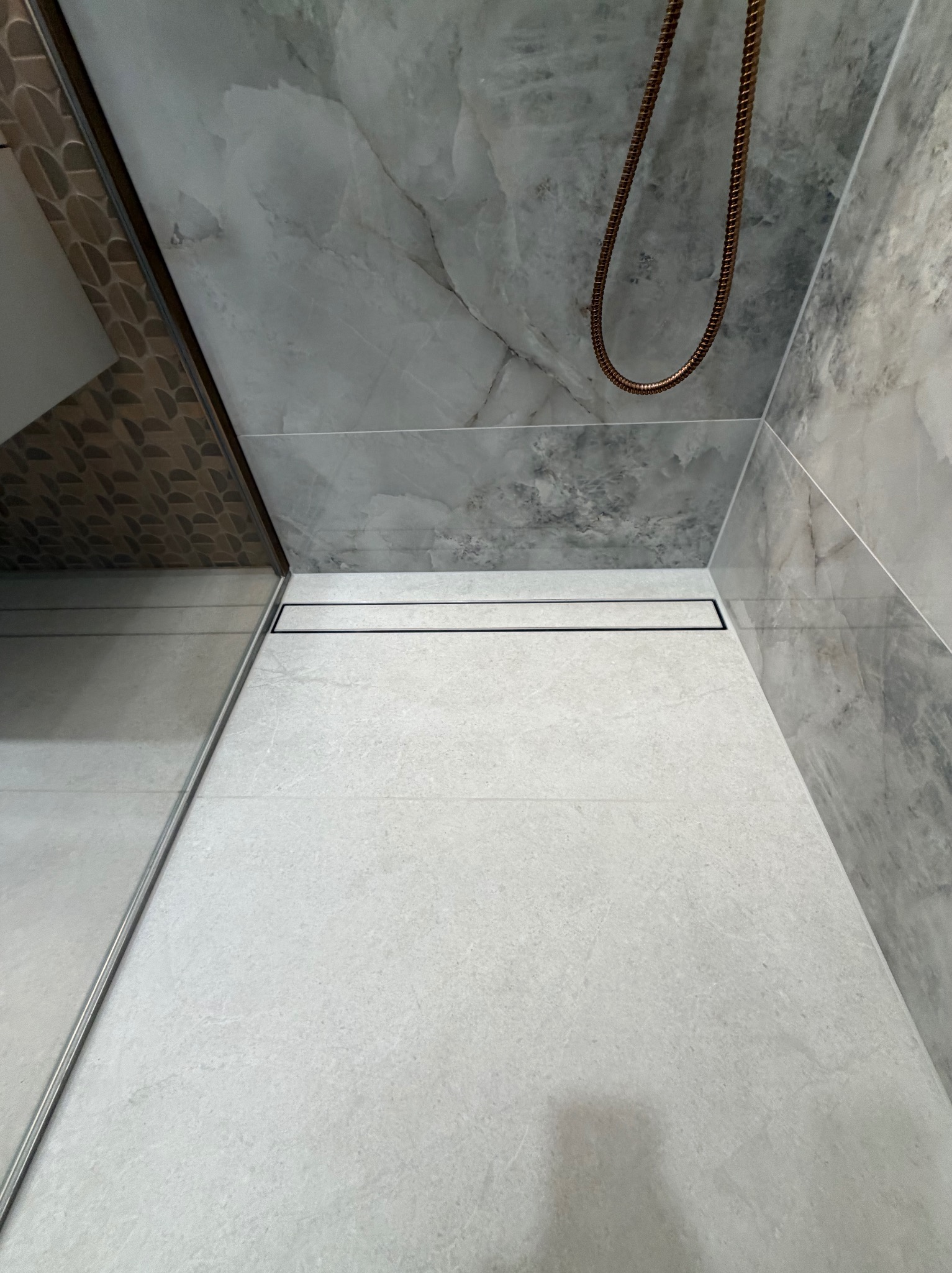 Nowoczesna kabina prysznicowa z odpływem liniowym i marmurowymi płytkami w odcieniach szarości. Widoczny fragment deszczownicy w kolorze miedzianym.