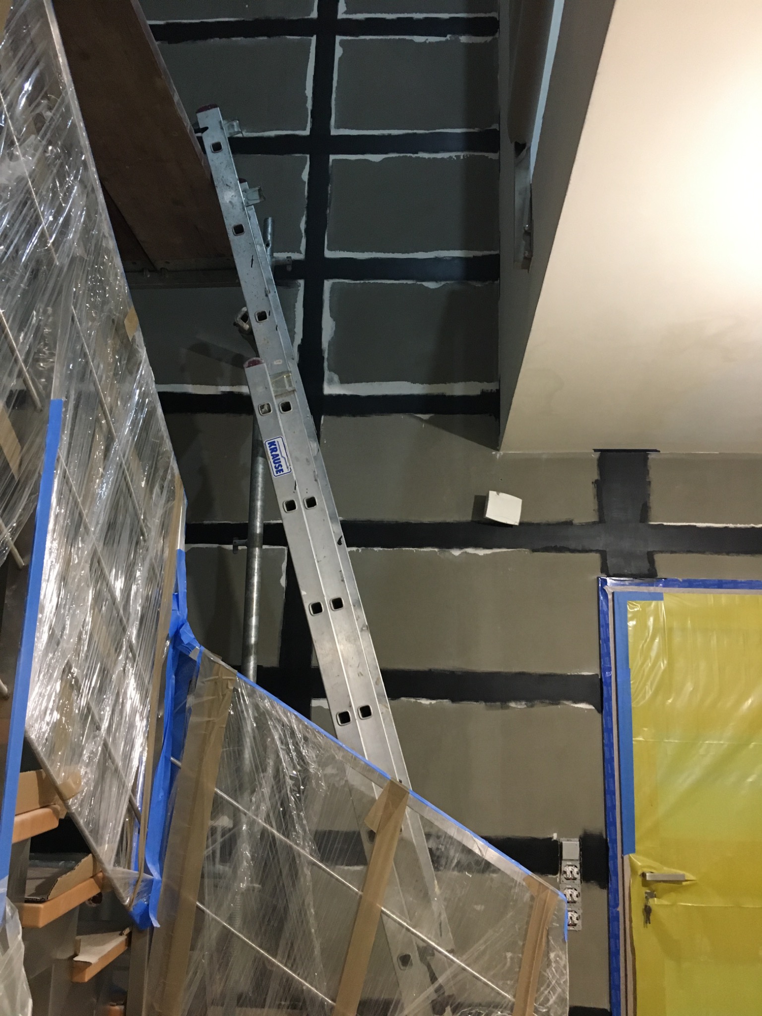 Wnętrze w trakcie prac wykończeniowych: ściany z widocznymi spoinami, aluminiowa drabina Krause oparta o ścianę, zabezpieczone folią schody z balustradą oraz drzwi osłonięte żółtą folią ochronną.