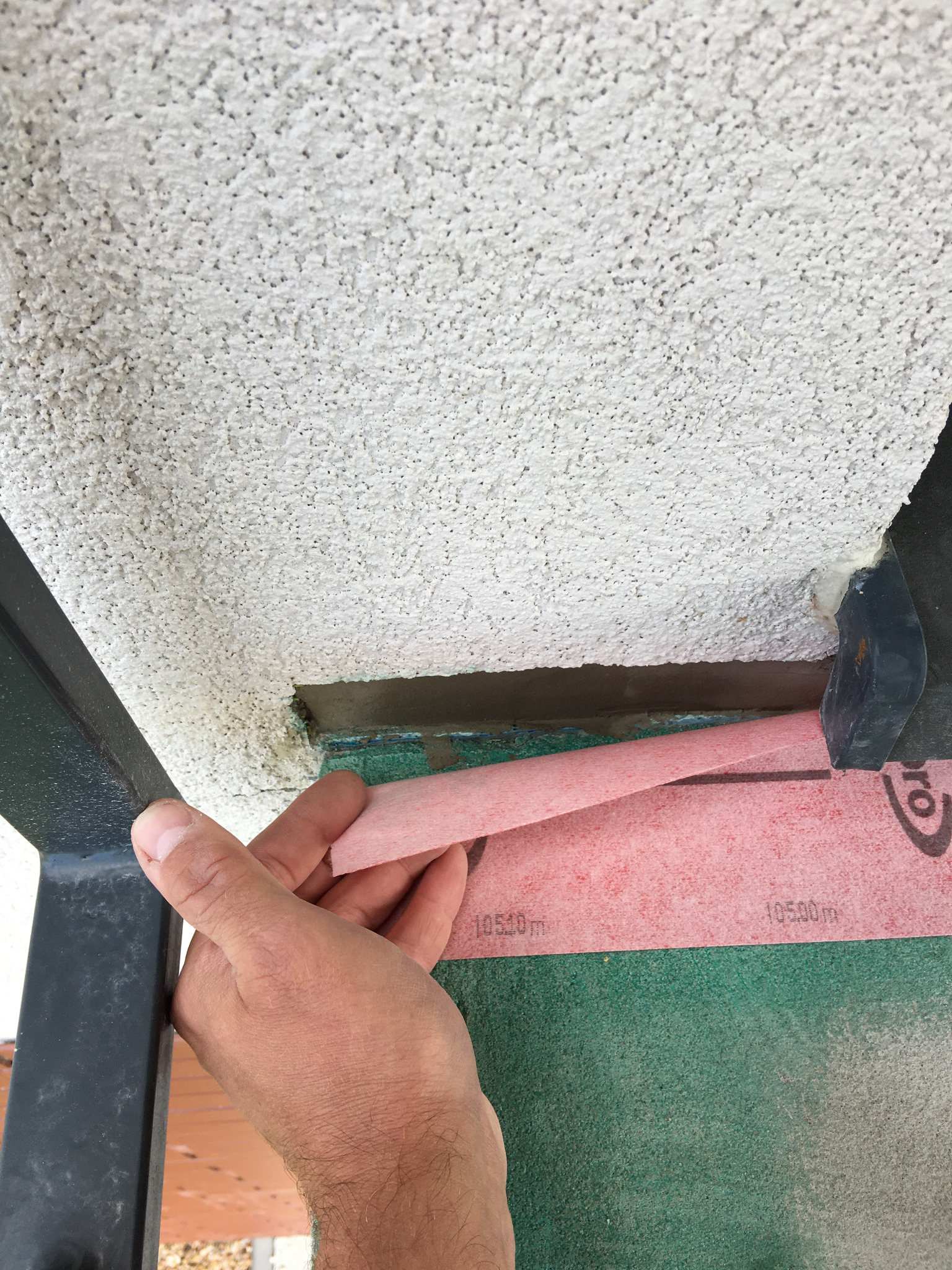 Prace wykończeniowe balkonu: Ręka przytrzymuje element konstrukcyjny balkonu, obok widoczna warstwa izolacyjna i młotek, sufit z fakturą baranka.