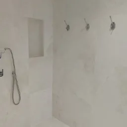 Wykończona kabina prysznicowa z beżowymi płytkami imitującymi marmur, wnęką na kosmetyki i trzema chromowanymi haczykami na ścianie.